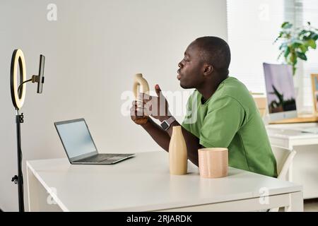Young African American man with handmade vase in hand talking to online audience while looking in smartphone camera during livestream Stock Photo