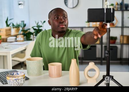 African American guy starting recording or livestream while sitting by desk with handmade items and pressing button on smartphone screen Stock Photo