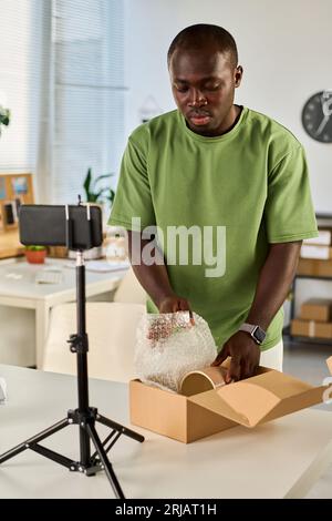 Young African American blogger unpacking cardboard box with vase or bowl during livestream while standing in front of smartphone Stock Photo