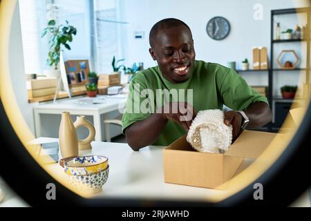 Happy young African American man putting wrapped handmade ceramic vase or some other earthenware item into cardboard box before sending Stock Photo