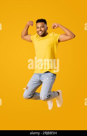 Full length portrait of overjoyed athletic indian guy jumping high and showing biceps, young man in casual wear isolated on yellow color background raising fists up in victory sign Stock Photo