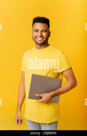 Smiling young Indian male freelancer stands in studio isolated on yellow and carrying trendy laptop. Cheerful arab guy in casual t-shirt looks at camera, holding closed laptop, student Stock Photo