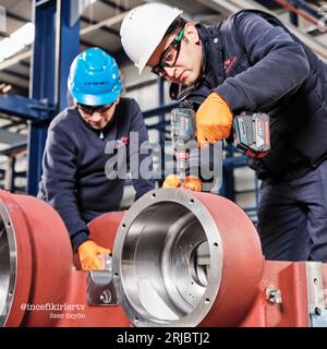 two men working on a metal pipe and using a drill in a factory while wearing work clothes, safety glasses, and helmets. Stock Photo