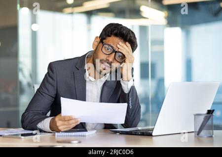 Upset and disappointed businessman financier investor looking at camera, man holding papers reports and contracts, unhappy with bank achievement results inside office with laptop. Stock Photo
