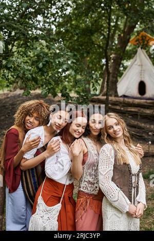 happy interracial women in boho outfits hugging while standing in retreat center Stock Photo