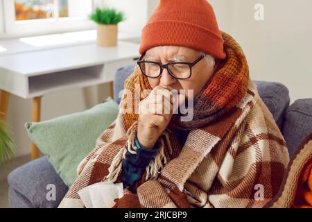 Sick senior man in hat wrapped in a blanket suffering from seasonal flu or cold at home. Stock Photo