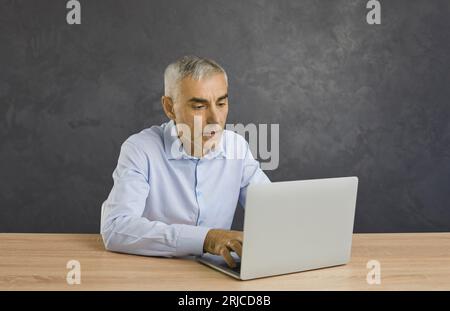 Portrait of serious and focused senior man typing on laptop sitting on gray background. Stock Photo