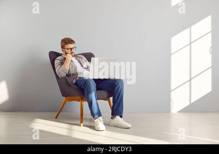 Young man sitting in armchair, working on his modern laptop computer, and thinking Stock Photo