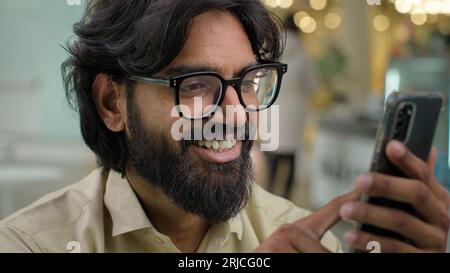 Smiling millennial Indian bearded guy businessman sit in cafe office worker in eyeglasses looking at smartphone screen using mobile phone app Stock Photo