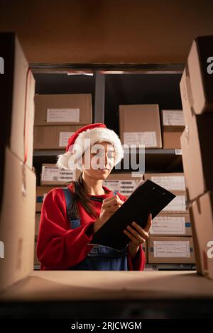 Female inventory manager checks stock, writing notes in the clipboard. Woman wearing Santa hat working in a warehouse storeroom with shelves full Stock Photo