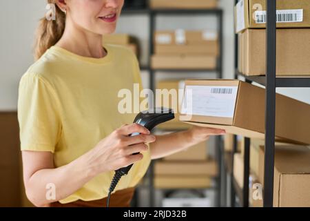 Cropped shot of young female worker of warehouse scanning qr codes on boxes while standing in front of shelves with packed online orders Stock Photo