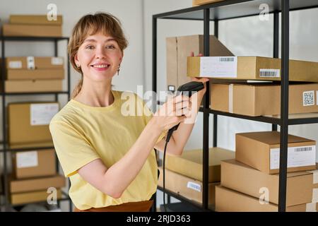Happy young female manager of marketplace with scanner looking at camera while checking qr codes on packed boxes with ordered goods Stock Photo