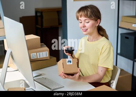 Young smiling woman scanning packed box with scanner while sitting by workplace in front of computer and checking qr code on package Stock Photo