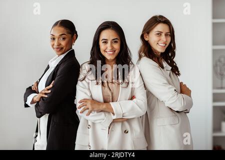 Business team. Three diverse female colleagues in formal outfits standing with folded arms in office, smiling at camera Stock Photo