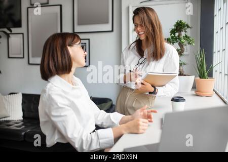 Cheerful female colleagues managers having conversation while working together in cozy office interior Stock Photo