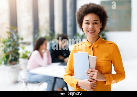Portrait of happy black lady university student posing with notepads while classmates studying on background in audience Stock Photo