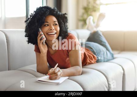 Happy Black Woman Talking On Cellphone While Lying On Couch At Home Stock Photo
