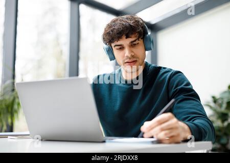 Remote studying. Concentrated european guy in headphones watching video course on laptop and taking notes Stock Photo