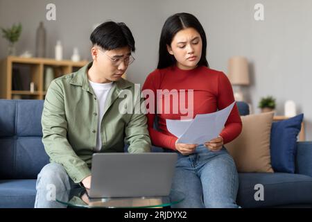 Stressed Korean Couple Calculating Budget On Laptop, Holding Papers Indoors Stock Photo