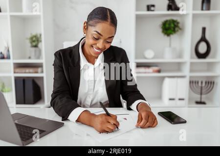 Happy black female business analyst writing on papers, checking data, charts and graphs, sitting at desk in light office Stock Photo