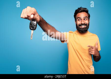 Excited indian man driver holding in hand new car key, pointing at it and smiling isolated on blue background Stock Photo