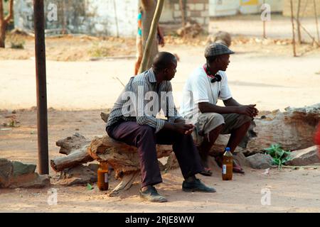 Two men are seen sitting on a fallen tree trunk at a shopping centre in Binga district. The two were consuming opaque  beer early in the morning. Zimbabwe. Stock Photo