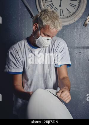 Focused adult blond haired tattooed male master grinding white handmade surfboard while shaping it with sandpaper in workshop Stock Photo