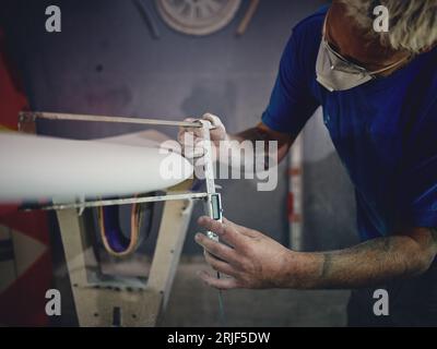 Crop attentive male shaper with blond hair and tattoos on arms in casual clothes and respirator measuring surfboard thickness with caliper during work Stock Photo