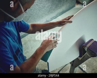 From above of crop anonymous male master grinding edges of surfboard with professional abrasive equipment in workshop Stock Photo