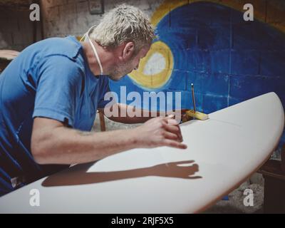 Side view of concentrated bearded male carpenter measuring thickness of edges of surfboard with caliper in workshop Stock Photo
