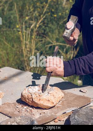 From above of crop anonymous craftsman cutting solid rock using sharp chisel and hammer while creating stone craft outside Stock Photo
