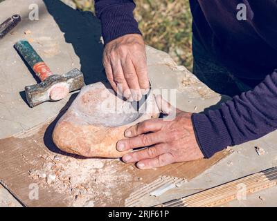 From above of crop unrecognizable elderly artisan using sandpaper for grinding solid rock while creating handmade stone bowl in sunlight Stock Photo