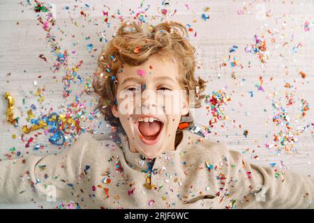 Top view of delighted preteen little boy with curly blond hair lying on laminated floor and screaming wow while enjoying falling confetti and sparkles Stock Photo