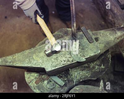 From above of crop anonymous blacksmith in protective gloves using hammer and iron sickle while working on anvil in workshop Stock Photo
