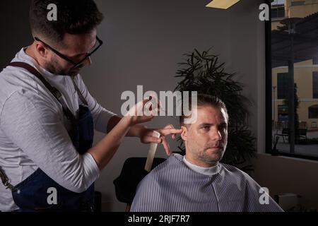 Focused skilled male hairdresser in apron and eyeglasses cutting hair of client with scissors while working in professional light barbershop Stock Photo