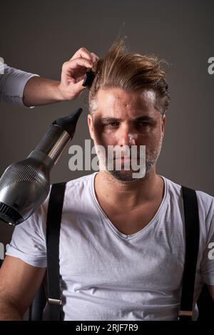 Confident bearded adult male in casual clothes and unrecognizable barber drying hair of client and combing it with hairbrush Stock Photo