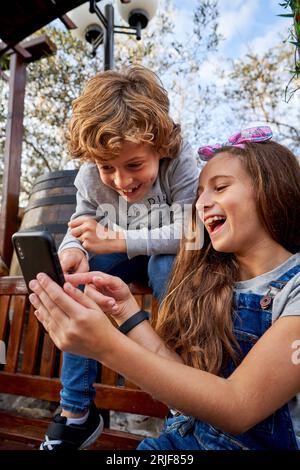 Cheerful siblings in casual wear smiling and watching video on mobile phone while sitting together on wooden bench in park on summer day Stock Photo