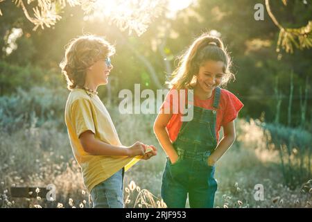 Happy boy and girl with hands in pockets telling jokes while standing in field with dry grass together on sunny summer day in countryside Stock Photo