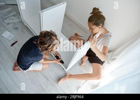 From above full body of concentrated barefoot woman and boy sitting on floor near window in living room and assembling wardrobe with screwdriver Stock Photo