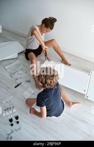 High angle of concentrated woman tightening screws with screwdriver while preteen boy reading instruction and helping in living room at home Stock Photo