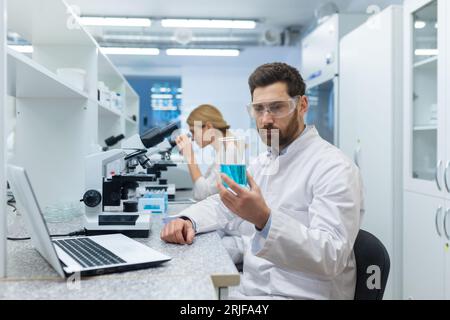 A young male scientist works in a laboratory. He sits at the work table behind the microscope, spits out and examines the blue substance in the flask. A female colleague works nearby. Stock Photo