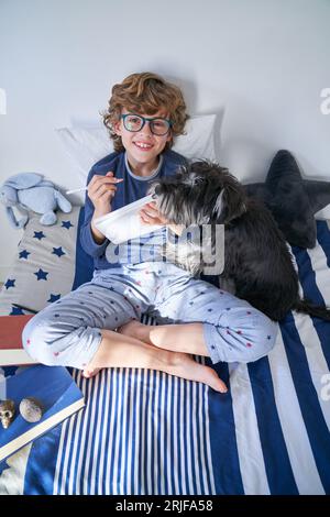 From above of cute curly haired child in pajama looking at camera and taking notes while sitting on bed together with dog and books and doing homework Stock Photo