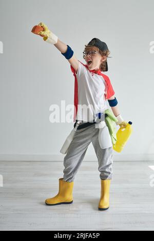 Full body of excited boy standing near studio gray wall having fun and raising hand with soap while looking away and getting ready for household chore Stock Photo