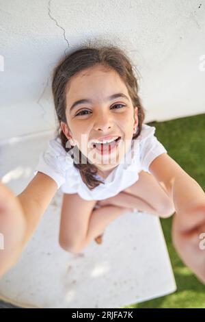 Full body top view of smiling preteen girl in white t shirt sitting near building wall with crossed legs and raising hands to camera Stock Photo