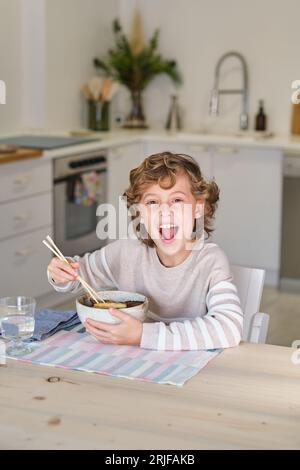 Excited child in casual clothes having fun while eating delicious noodles with chopsticks in kitchen at home Stock Photo