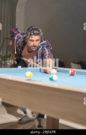 Serious middle aged bearded man with stylish haircut playing billiards in living room with cocktails and sofa on background Stock Photo