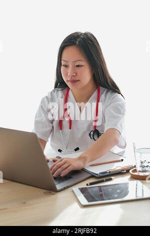 Concentrated Asian female doctor in medical uniform typing on netbook while working at table with tablet in modern light medical office Stock Photo