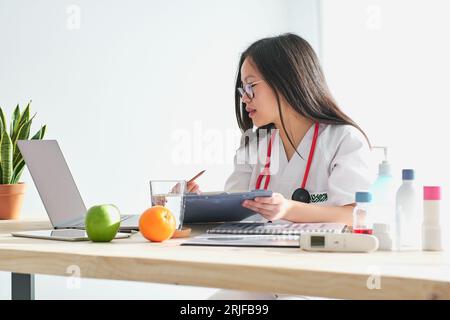 Side view of concentrated Asian female therapist in medical gown surfing netbook while working with documents at table in modern light clinic Stock Photo