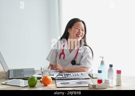 Cheerful Asian female medic in white uniform sitting at table with laptop and documents while working in modern light clinic Stock Photo