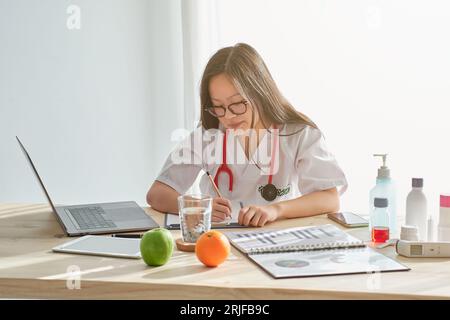 Concentrated Asian female physician in uniform taking notes on clipboard while working at table with laptop in modern light clinic Stock Photo